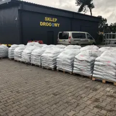 Palety z workami materiału budowlanego przed budynkiem sklepu drogowego.