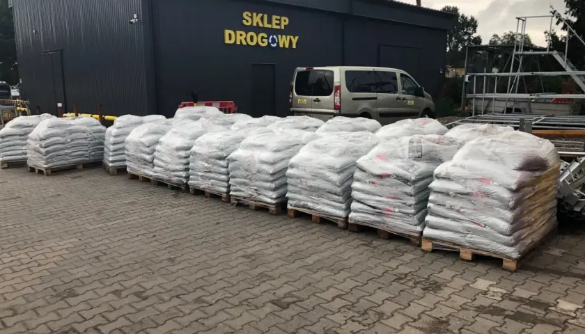 Palety z workami materiału budowlanego przed budynkiem sklepu drogowego.